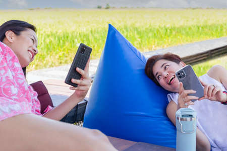 Two woman friends lying on cushion and holding smart phone with nature view background.の写真素材