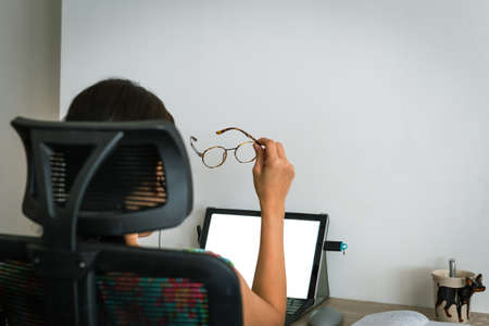 Asian businesswoman holding eyeglasses while working overtime on laptop at home.