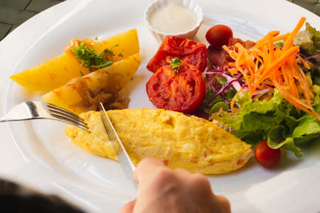 Woman hand holding fork and knife eating breakfast with omelet potato, tomatoes and vegetable salad on table.の写真素材