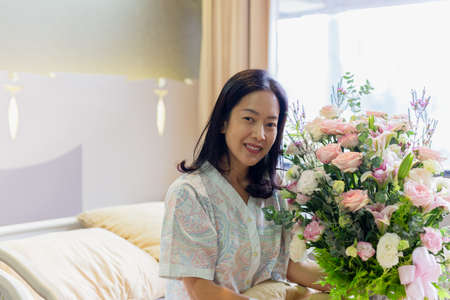 Woman patient smiling and holding a flower bouquet sitting on hospital bed.の写真素材