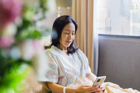 Asian woman patient smiling while sit on bed making video call on cell phone in hospital ward.の写真素材