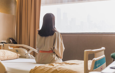 Back view of patient woman sitting on bed in hospital ward looking out at window.の写真素材