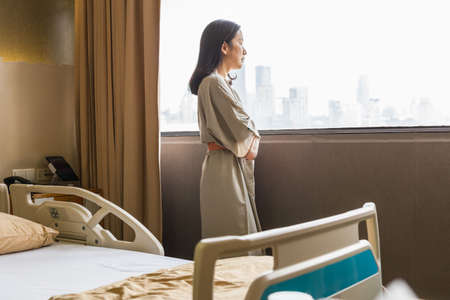 Depressed asian woman patient standing is hospital ward look out window.