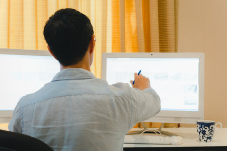 Back View of man working on Pc computer sitting at desk at home office.