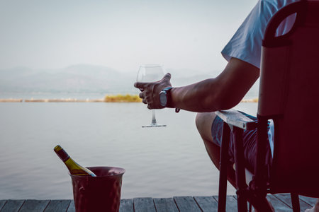 Man sitting on a chair with hand holding glass of red wine by the lake.の写真素材