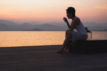 Silhouette woman holding cup coffee sitting by the lake watch sunrise.の写真素材