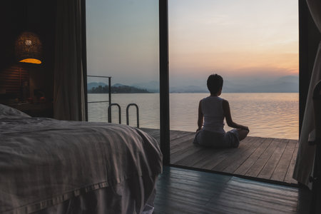 Woman sitting meditation on a wooden balcony with a beautiful sunrise of a mountain lake.の写真素材