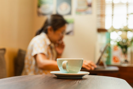 Selected focus cup of coffee on table with woman working on laptop in blur backgroundの写真素材