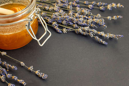 Open glass jar of liquid honey and honey dipper inside and bunch of dry lavender over dark surface.