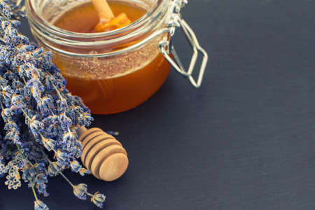 Open glass jar of liquid honey with honeycomb and honey dipper inside and bunch of dry lavender over dark surface.の写真素材
