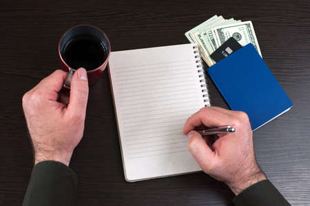 Top view of businessman writing on blank notebook. Empty notebook, banknotes, passport and coffee cup on dark wooden table. Copy spaceの写真素材