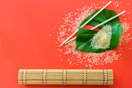 Pile of rice with chopsticks, bamboo mat and leaf on red background. Top view.の写真素材