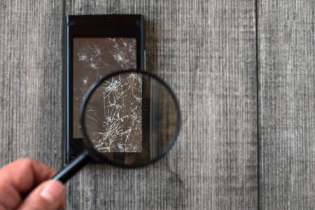 Broken Smart phone and magnifier in hand on gray wooden background