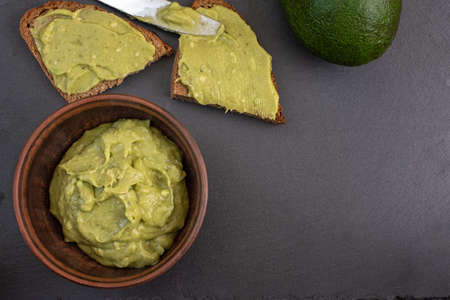 Fresh avocado cream in brown bowl with bread slices on black background. Top view. With copy space.
