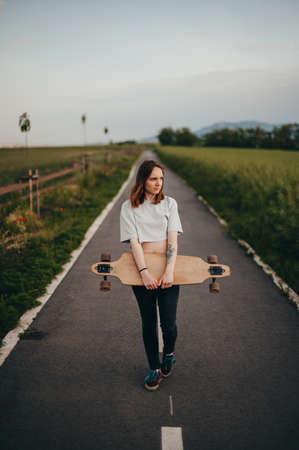 Vertical poster. The girl holds a longboard in her hand on a dirt road and looks awayの写真素材