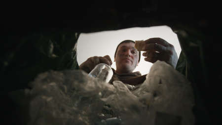 The homeless man digs in the trash. Bottom view from the trash bin. Homeless man looking for food in a basketの写真素材