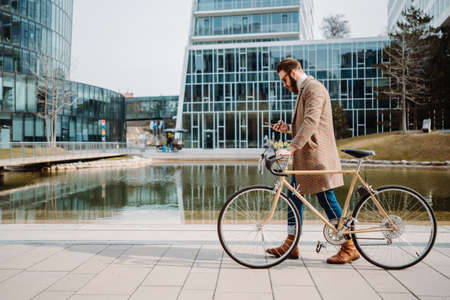 Young hipster businessman walking with bicycle. Using smartphone. Eco friendly transport