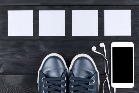 Overhead view of sports shoes by mobile phone with isolated screen and in-ear headphones and white empty blank sheets on black wooden table, floor. Empty space for text, copy space, empty blank screenの写真素材