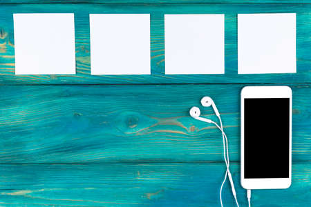 Overhead view of mobile phone with isolated screen and in-ear headphones and white empty blank sheets on black wooden table, floor. Empty space for text, copy space, empty blank screenの写真素材