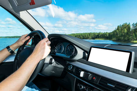 Male hands holding car steering wheel. Hands on steering wheel of a car driving near the lake. Man driving a car inside cabin. Multimedia system isolated white blank screen. Copy spaceの写真素材