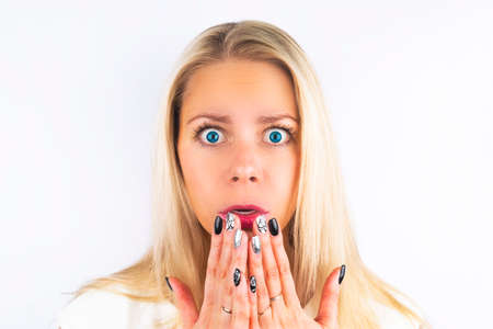Surprised blond girl with blue eyes screams and closes mouth with her hands from surprise. Presenting your product. Isolated on white background. Expressive facial expressions. Surprised happy young womanの写真素材