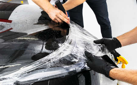Car wrapping specialist putting vinyl foil or film on car. Protective film on the car. Applying a protective film to the car with tools for work. Car detailing. Transparent film. Car paint protection. Trimming.の写真素材