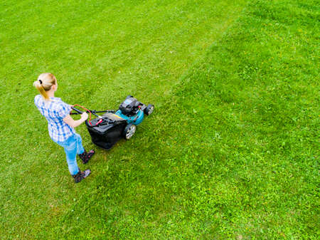 Beautiful girl cuts the lawn. Mowing lawns. Aerial view beautiful woman lawn mower on green grass. Mower grass equipment. Mowing gardener care work tool. Close up view. Aerial lawn mowingの写真素材