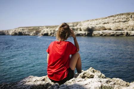 Young woman sitting on the top of rock and looking at the seashore and mountains at colorful sunset in summer. Landscape with girl, sea and mountain. Russia, Crimea.の写真素材