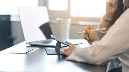 Two young business women sitting at table. First woman holding stylus pen with digital tablet screen. close up side view.の写真素材