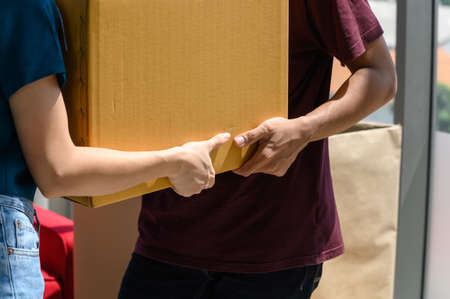 close up to hand of young asian couple when they carrying big cardboard box while relocation into new home on moving day, celebrating moving to new home conceptの写真素材