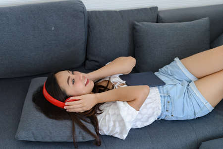 Portrait of beautiful young Asian woman wearing headphones to listening music while relaxing on sofa...