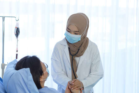 Muslim female doctor holding hand of the elderly patient sitting on the bed at hospital, takes care...