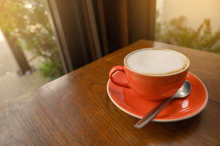 Orange cup of hot cappuccino coffee on wooden table within cafe near glass window, modern lifestyle concept.の写真素材