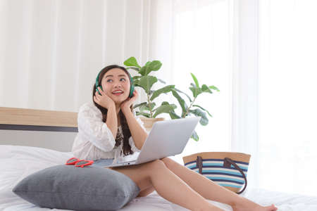 Beautiful young Asian woman listening music in headphones relaxing while using laptop on bed in...