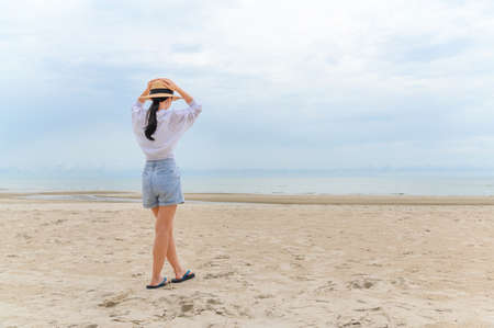 Outdoor summer portrait of young asian woman in straw hat looking to the sea at tropical beach, enjoy freedom time, fresh air and leisure travel vacationの写真素材