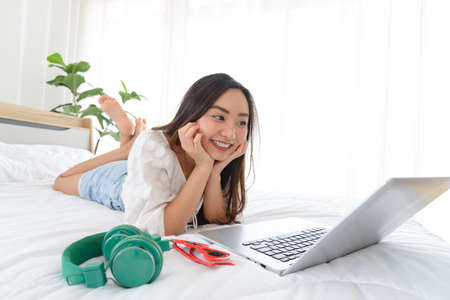 Beautiful young Asian woman relaxing and using a lapto on her bed.