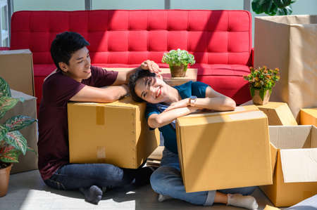 Young asian couple smiling, relaxing and resting on the floor after unpacking cardboard boxes in new...