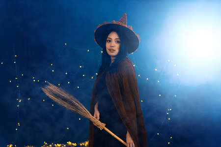 Portrait of beautiful young woman in witch halloween costume wear witches hat holding broom over spooky dark magic background - Halloween party art designの写真素材