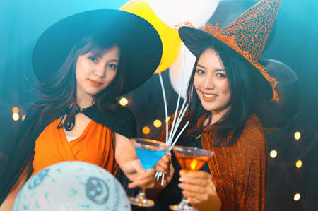 Two beautiful young woman in witch halloween costumes drinking cocktail and holding balloons in halloween party over dark magic background - Holiday party conceptの写真素材