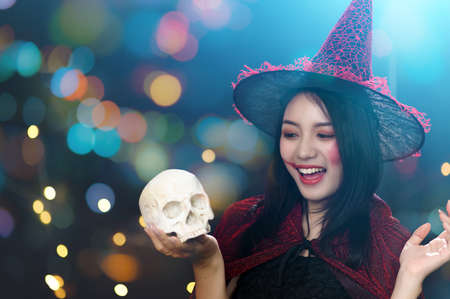 Portrait of beautiful young woman in witch halloween costume wear witches hat hold human skull over spooky dark magic background - Halloween party conceptの写真素材
