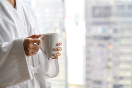 Side view of woman in bathrobe standing near the window while holding white cup of coffee and enjoy wonderful morning in the hotel, detaching from workの写真素材