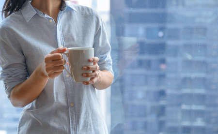 Close-up woman hands holding white cup of coffee or tea by the window with blurry of city view high building on background. Copy spaceの写真素材