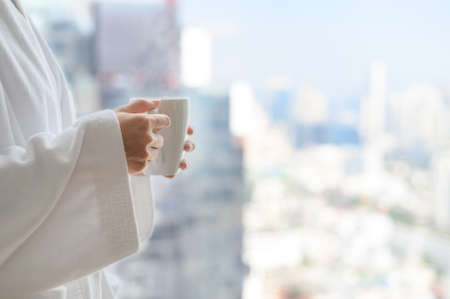 Side view of woman in bathrobe standing near the window while holding white cup of coffee and enjoy wonderful morning in the hotel, detaching from workの写真素材