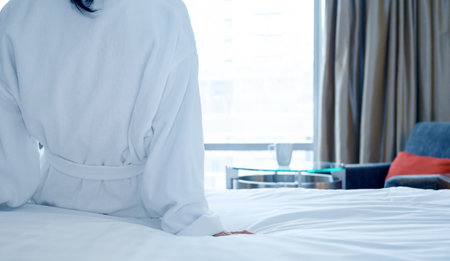 Rear view of woman in white bathrobe sitting on bed after waking up, looking at city view in the window. Copy spaceの写真素材