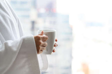 Side view of woman in bathrobe standing near the window while holding white cup of coffee and enjoy wonderful morning in the hotel, detaching from workの写真素材