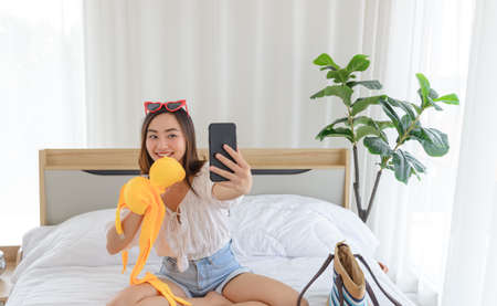 Young Asian woman show yellow	bikini while talking to calling with her friend in bedroom. Using smartphone taking selfie on front cameraの写真素材