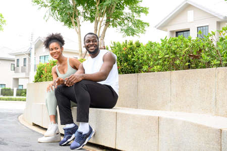 Happy young African American couple sitting after workout outdoors, wear sports clothes resting,...