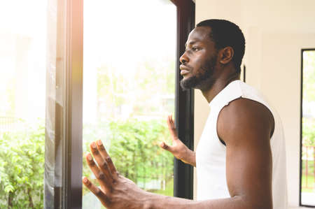 Portrait of pensive African American man standing, touching, and looking outside the window at homeの写真素材