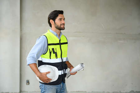 Portrait ofÂ young attractive construction man smiling bearded in vest and holding white helmet and blueprint while standing on construction site. Home building conceptの写真素材