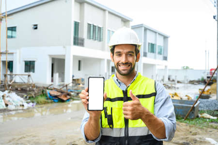 Young attractive construction man smiling bearded in vest with white helmet demonstrating blank smartphone and showing thumb up to the camera while standing on building construction site. White screen, mockupの写真素材